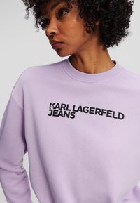 Light purple sweatshirt featuring a round neckline and long sleeves. Black "KARL LAGERFELD JEANS" logo printed prominently on the front.