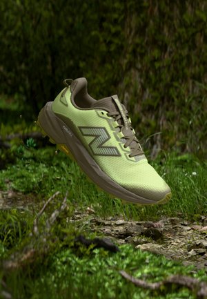 Zapatilla de trail running New Balance verde claro y marrón flotando sobre un suelo forestal rocoso y con hojas.