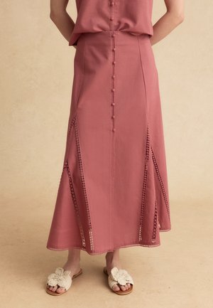 Donna che indossa una gonna midi rosa polvere con bottoni, pannelli in crochet e sandali crema con fiori all'uncinetto, in piedi davanti a uno sfondo semplice.