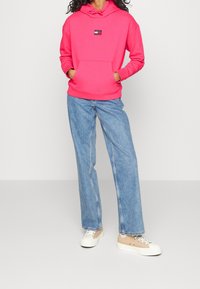 Persona che indossa un maglione a cappuccio Tommy Jeans di un vivace rosa, jeans a gamba larga di un blu chiaro e sneakers beige a bassa altezza, in piedi con le mani nella tasca del cappuccio.