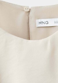 Encolure de vêtement beige avec une petite fermeture bouton et une étiquette blanche visible portant la marque MNG à l'intérieur au dos.