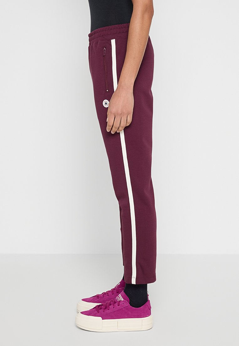 Los pantalones de chándal burdeos presentan una franja lateral blanca, cintura elástica y un bolsillo con cremallera. Se combinan con zapatos de color rosa brillante y calcetines negros.