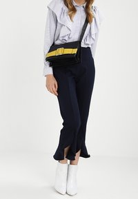 Donna che indossa una blusa a righe con volant, pantaloni navy a culotte con spacchi svasati, stivaletti bianchi alla caviglia e una borsa a tracolla nera con fibbia gialla.