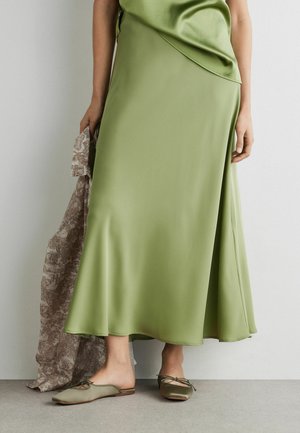 Kvinde iført en grøn satin midi-nederdel og matchende slip-on flade sko, der holder et brunt mønstret stof i venstre hånd.