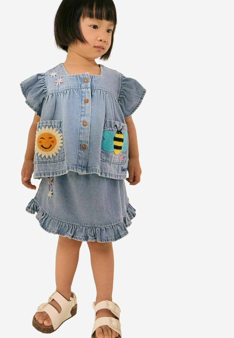 Jeanskleid mit kurzen Ärmeln, Rüschensaum, Sonne- und Bienenapplikationen an den Taschen, hellblaue Farbe, kombiniert mit weißen Sandalen.
