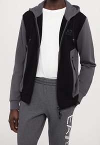 Svart och grå zip-up hoodie med huva, tillverkad av mjukt material; har sidfickor och en logotyp på bröstet. Matcher med gråa sweatpants.