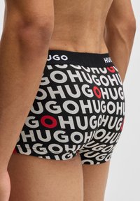 Bóxer negro con un logo repetido de "HUGO" en blanco y rojo, con una cinturilla negra que muestra el nombre de la marca de manera prominente.