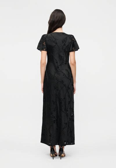 Γυναίκα με μακριά σκούρα μαλλιά, που φοράει μαύρο μακρύ φόρεμα με κεντήματα λουλουδιών, κοντά φουσκωτά μανίκια και μαύρα σανδάλια με ψηλό τακούνι, στέκεται με την πλάτη.