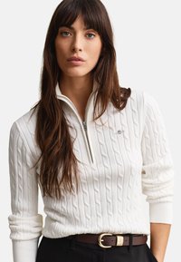 Γυναίκα με μακριά καστανά μαλλιά, φορεμένη με λευκό πουλόβερ με ορειβατική πλέξη και φερμουάρ μέχρι τη μέση και σκούρο ζωνάρι με χρυσή αγκράφα.