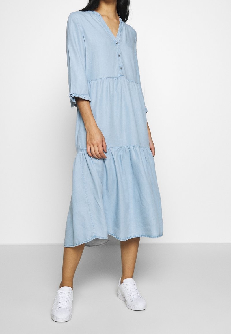 Femme debout portant une robe midi bleu clair avec des boutons et manches 3/4, associée à des baskets blanches sur fond uni.