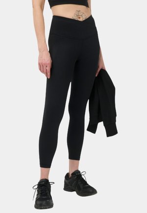 Persoon draagt zwarte hoge taille leggings, zwarte sneakers en een cropped top, en houdt in één hand een zwart jasje vast.