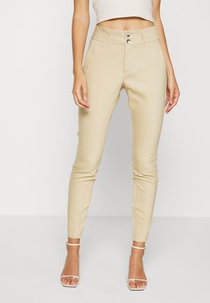 Femme portant un pantalon beige ajusté à taille double boutonnage et des escarpins blancs ouverts, debout devant un fond clair uni.