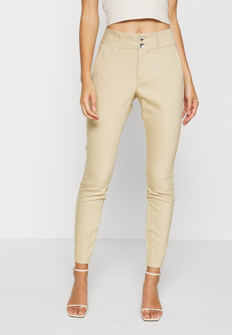 Femme portant un pantalon beige ajusté à taille double boutonnage et des escarpins blancs ouverts, debout devant un fond clair uni.