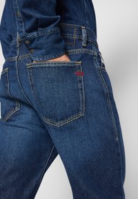 Dark blue denim jeans with a straight cut, featuring yellow stitching, two back pockets, and a small red logo on one pocket.