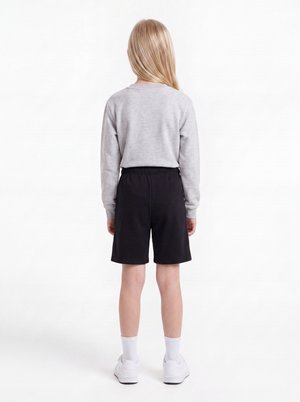 Barn med langt blondt hår står med ryggen til iført en lysegrå sweatshirt, sorte shorts, hvide sokker og hvide sneakers på hvid baggrund.