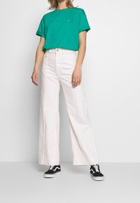 T-shirt bleu sarcelle à col rond avec des manches retroussées, associé à un pantalon blanc taille haute à jambes larges. Des baskets noires complètent la tenue.