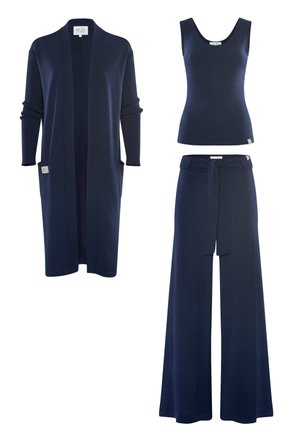 Junge Frau mit roten Haaren steht barfuß, trägt ein dunkelblaues Tanktop, weit geschnittene Hosen mit Gürtel und eine lange Strickjacke vor einem einfarbigen Hintergrund.