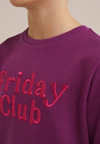 Fialová mikina s vyšitým textom "Friday Club" v ružovej farbe. Hladký rebrovaný golier s textúrovaným povrchom. Detailný pohľad.