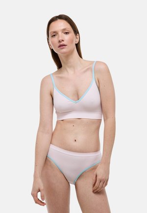 Femme portant un ensemble de lingerie rose clair avec des bordures bleues, debout devant un fond gris clair uni, regardant légèrement vers l'avant.