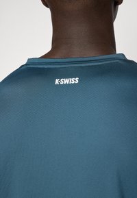 Tealová sportovní košile K-Swiss viděná ze zadní strany, s kulatým výstřihem a texturovanou tkaninou s bílým logem na výstřihu.