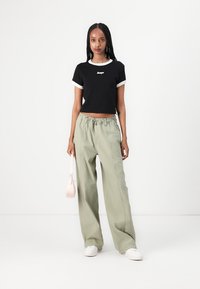 Svart cropped t-skjorte med hvit kant og logo, paret med løse, lysegrønne bukser og hvite sneakers. Holder en liten rosa håndveske.
