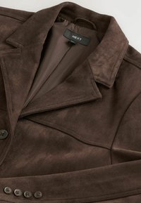 Veste en suède synthétique brun avec col cranté, fermetures à bouton et doublure intérieure lisse. Le tissu présente une texture douce.