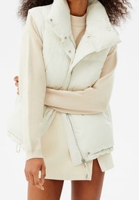 Gilet rembourré gris clair avec un col haut, des boutons-pression et une fermeture éclair. Porté par-dessus un pull beige et une jupe crème, mettant en valeur un look superposé.