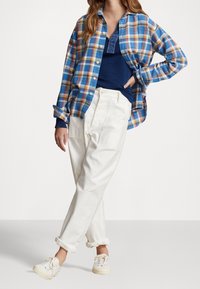 Rutig flanellskjorta i blått, gult och rött; lager över en blå tröja. Vita byxor med avslappnad passform och vita sneakers. Vänd upp ärmsluten.