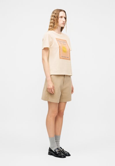 Γυναίκα με κυματιστά μαλλιά που φοράει μπεζ γραφικό t-shirt, καφέ σορτς, γκρι κάλτσες και μαύρα μοκασίνια, στέκεται μπροστά σε απλό λευκό φόντο.