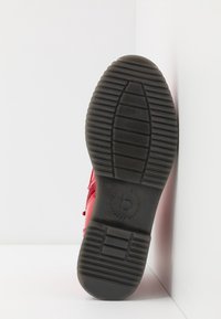 Semelle d'une basket rouge avec une semelle en caoutchouc noire présentant des rainures horizontales, des motifs rectangulaires et un logo circulaire avec la lettre "b" au centre.