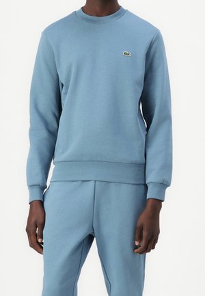 Man draagt een lichtblauwe trui en bijpassende broek met een klein groen krokodillenlogo op de linkerborst.