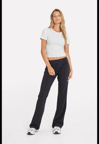 Junge Frau steht, trägt ein weißes, kurzes T-Shirt, schwarze, hochtaillierte, weite Hosen und weiße Turnschuhe vor einem schlichten weißen Hintergrund.
