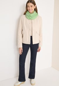 Femme souriante debout portant une veste beige boutonnée, un jean évasé foncé, des baskets blanches et une écharpe verte à l'intérieur contre des murs blancs.