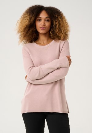 Maglione a coste rosa chiaro con maniche lunghe, vestibilità rilassata e fessure laterali, abbinato a pantaloni neri aderenti. Dettaglio di texture sottile su tutta la superficie.