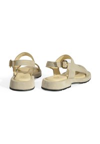 Beige suède sandalen met een achterband en een gouden gesp, een gevoerde binnenzool en een dikke zwarte buitenzool. Ontworpen met open zijkanten voor ademend vermogen.