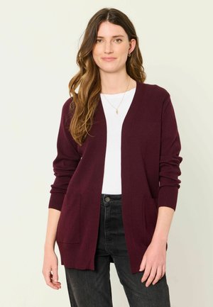 Woman with long brown hair wearing a burgundy cardigan over a white shirt and black jeans against a plain light background.