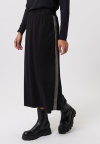 Zwarte rok met elastische tailleband en zijstrepen. Gemaakt van zacht materiaal, met een vloeiend ontwerp en gaat tot halverwege de kuit. Gecombineerd met zwarte laarzen.