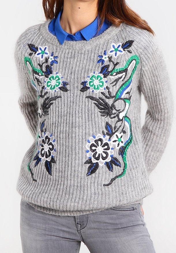 Pull gris en maille avec broderie florale verte, bleue, noire et blanche porté sur une chemise bleue à col et associé à un jean gris.