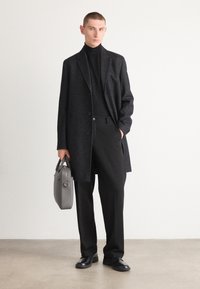 Cappotto nero con tessuto testurizzato, sovrapposto a un dolcevita nero, abbinato a pantaloni neri. Tiene una borsa grigio chiaro. Sfondo neutro.