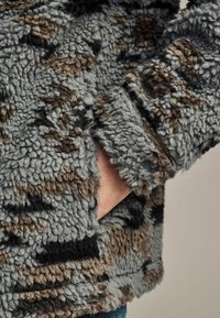 Fleecejacke in Grau mit braunen und schwarzen Mustern; verfügt über eine weiche, strukturierte Oberfläche und eine Seitentasche.