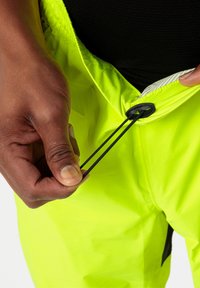 Pantaloni da atletica di un vivace giallo, con una vita elastica e un cordino nero. In primo piano, una mano che regola il cordino in vita.