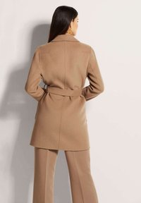 Beige wollen blend jas met een riem in de taille, getailleerd ontwerp, geknipte kraag en twee zijzakken. De broek is ook beige en op maat gemaakt.