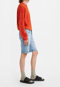 Camisa naranja de manga larga, pantalones cortos de mezclilla azul claro, calcetines de patrón oliva y sandalias negras tipo slide. Atuendo sencillo y casual con un corte holgado.