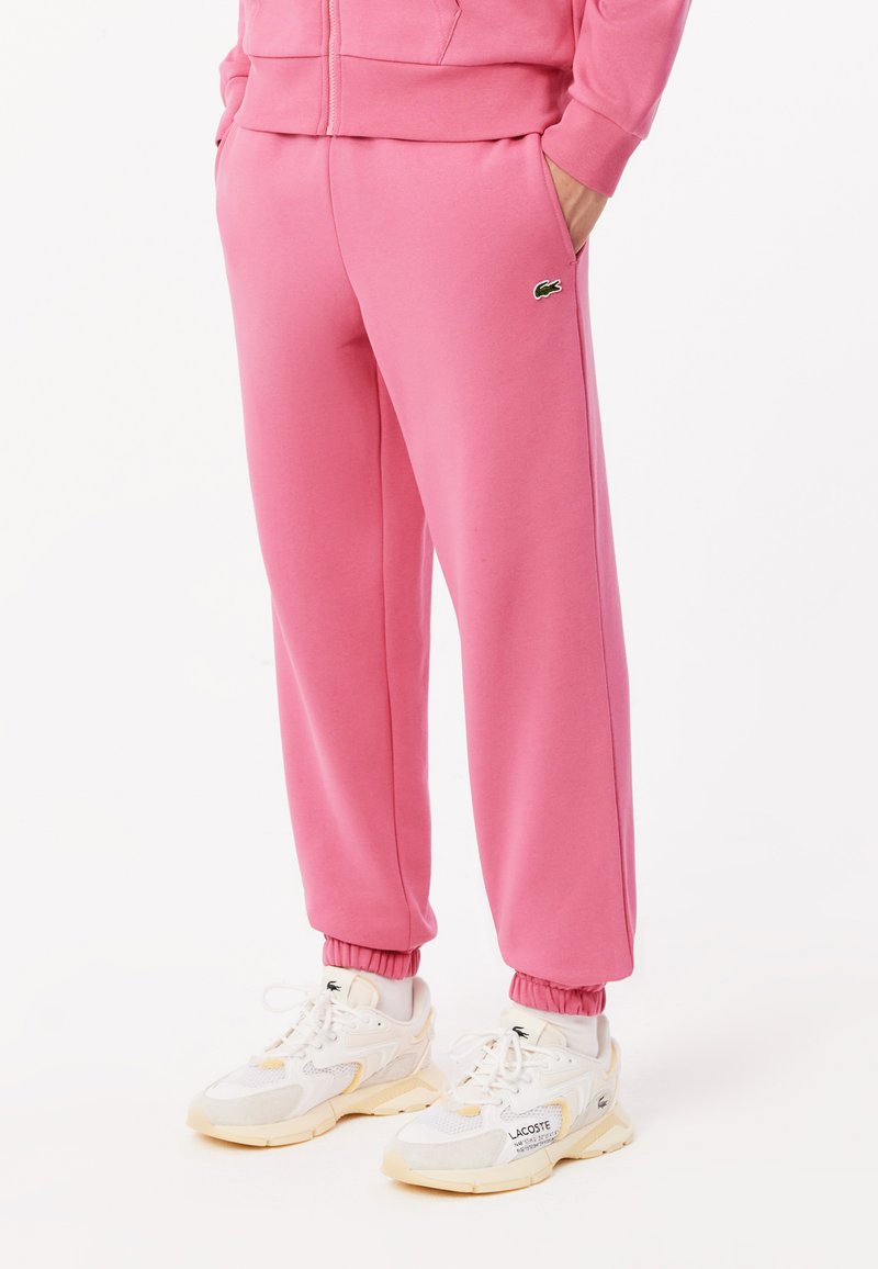 Pantalons de sport roses en tissu doux, avec des poignets élastiques et des poches latérales. Accompagnés de baskets blanches, affichant un détail de marque sur le côté.