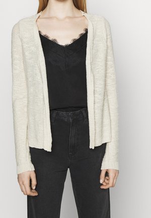 Cardigan aperto in maglia color avorio con una finitura texturizzata; abbinato a un top in pizzo nero e jeans scuri.