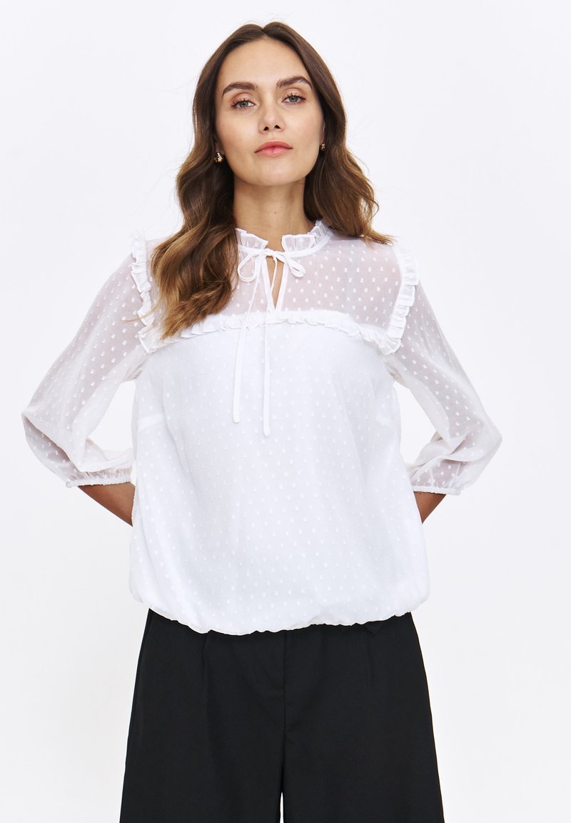 Biała bluzka w kropki, wykonana z przezroczystego materiału, z falbaniastym dekoltem i rękawami oraz wiązaniem ze ściągaczem przy kołnierzu. Luźny krój.