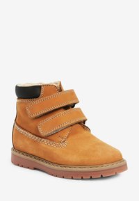 Bottine en suède tan avec des sangles Velcro doubles, accent noir au col, doublure en fourrure douce et semelle en caoutchouc texturé.