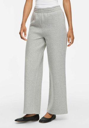 Personne portant un pantalon de jogging gris clair à jambes larges et des chaussures plates noires, debout devant un fond blanc.