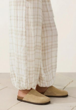 Person iført løse, ternede hvide bukser stoppet ned i beige slip-on ruskindsklumpfødder stående på flisebelagt gulv.