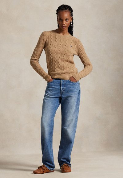 Femme debout, les mains dans les poches, portant un pull beige à torsades, un jean bleu et des mocassins marron sur un fond uni.
